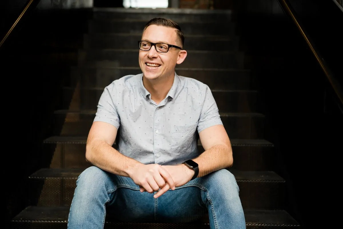Smiling man in a gray shirt seated on dark steps, environmental portrait